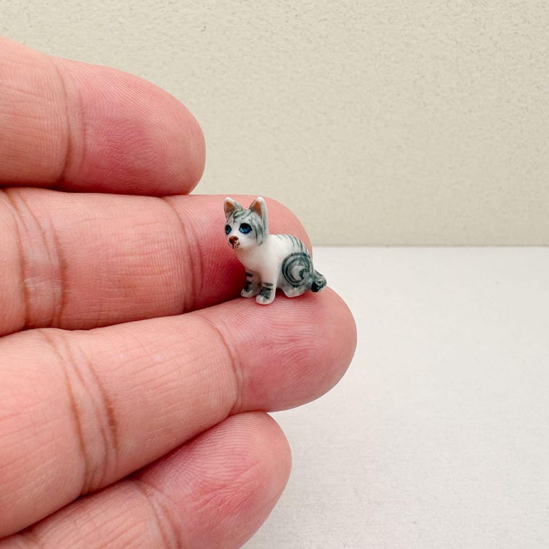 May include: A miniature ceramic cat figurine. The cat is white with gray stripes and blue eyes. The small cat is sitting down with its tail curled. The figurine is held in a person's hand.