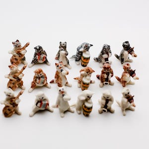 May include: A collection of 18 ceramic cat figurines playing musical instruments. The cats are painted in various shades of white, gray, brown, and black. Some cats are playing the violin, guitar, drums, and clarinet.