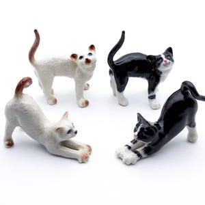 Könnte beinhalten: Vier Keramik-Katzenfiguren in verschiedenen Posen. Zwei sind weiß mit braunen Markierungen, eine ist schwarz mit weißen Markierungen und eine ist schwarz mit weißen Markierungen. Die Figuren sind alle etwa 7,5 cm hoch.