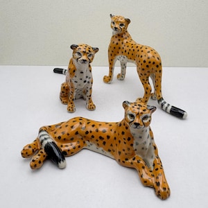 May include: Three ceramic cheetah figurines. One is lying down, another is sitting, and the third is standing. The cheetahs have orange bodies with black spots, white bellies, and black and white striped tails. The figurines are decorative.