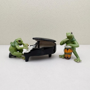 May include: Two ceramic frog figurines. One frog plays a black piano with white keys, while the other frog plays a small drum with orange and yellow patterns. The frogs are green and the piano is glossy.