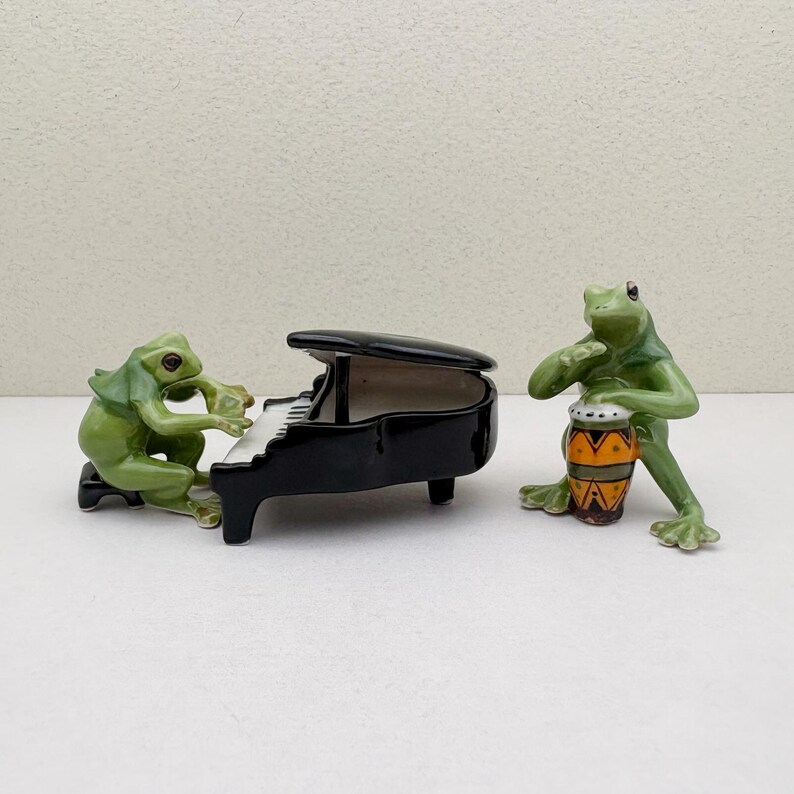 May include: Two ceramic frog figurines. One frog plays a black piano with white keys, while the other frog plays a small drum with orange and yellow patterns. The frogs are green and the piano is glossy.