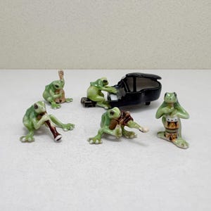 May include: A set of five small ceramic frog figurines arranged as a musical ensemble. One frog plays a black grand piano, while the others play a flute, a cello, an accordion, and a drum. Each frog is light green with detailed painted features.