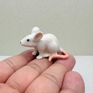 May include: A small, white ceramic mouse figurine with a pink tail. The mouse has black eyes and is posed in a crawling position. The figurine is held in a person's hand.