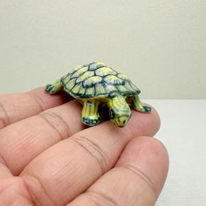 May include: A miniature ceramic turtle figurine. The turtle has a yellow and green shell with a detailed pattern. Its head and legs are also green, and it appears to be crawling. The figurine is small and detailed.
