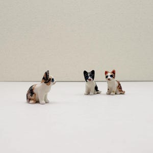 May include: Three small ceramic cat figurines. The cats are white with brown, black, and orange markings. The figurines are detailed and realistic. The cats are in a sitting position, arranged in a row on a white surface.