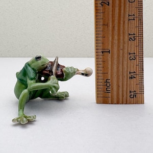 May include: A green frog figurine playing a brown violin. The frog is wearing a green jacket. The figurine is approximately 1 inch tall.