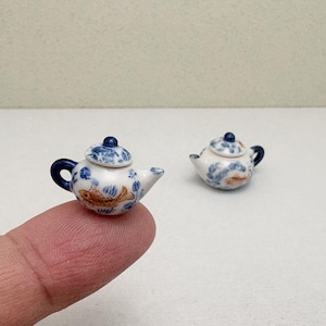 May include: Two miniature ceramic teapots. The teapots are white with blue and gold floral and fish designs. One teapot is held on a finger. The teapots have blue handles and lids.
