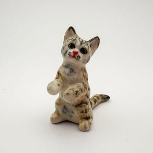 May include: A ceramic cat figurine in a standing position. The cat is patterned with brown, tan, and white speckles. It has a red nose, black eyes, and a striped tail. The cat is approximately 7.6 cm tall.