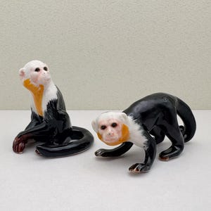 Pode incluir: Duas estatuetas de macaco de cerâmica. Uma está sentada com as pernas cruzadas e a outra está de pé em quatro patas. Ambos os macacos são pretos com rostos brancos e detalhes amarelos.