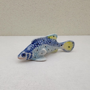 Op de afbeelding: Een keramieken vissenbeeldje in blauw, wit en geel. De vis heeft zwarte stippen en een rode mond. Het beeldje is een decoratief item, mogelijk voor een plank of tafel.