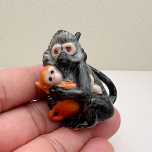 May include: A small ceramic figurine of a black and gray monkey holding a smaller orange monkey figurine. The monkey has white fur around its eyes.
