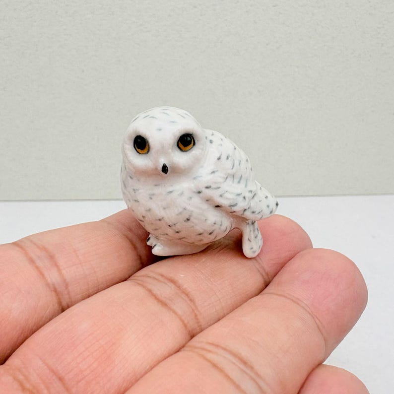 K&ouml;nnte beinhalten: Eine kleine, wei&szlig;e Eulenfigur mit schwarzen Sprenkeln und gro&szlig;en, bernsteinfarbenen Augen. Die Eule sitzt auf einer Hand und zeigt ihre detaillierten Merkmale und ihr realistisches Design. Die Figur ist eine Miniatur eines Schnee-Eulen.