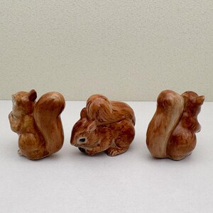 Peut inclure: Trois figurines de l'&eacute;cureuil en c&eacute;ramique dans diff&eacute;rentes poses. Les &eacute;cureuils sont de couleur brun chaud avec des textures de fourrure d&eacute;taill&eacute;es et de grandes queues touffues. Les figurines sont sur fond blanc.