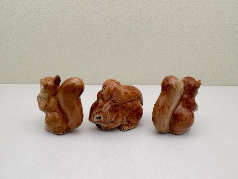 Peut inclure: Trois figurines de l'&eacute;cureuil en c&eacute;ramique dans diff&eacute;rentes poses. Les &eacute;cureuils sont de couleur brun chaud avec des textures de fourrure d&eacute;taill&eacute;es et de grandes queues touffues. Les figurines sont sur fond blanc.