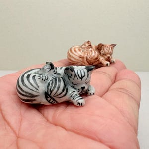 May include: Two miniature ceramic cat figurines, one featuring a white body with black stripes and the other a tan body with brown stripes. Each figurine depicts a larger cat curled up with a smaller kitten resting on its back, both with closed eyes.