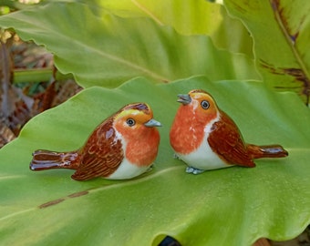 May include: Two ceramic bird ornaments with brown, orange, and white detailing. The birds are positioned on a large green leaf. One bird has its beak open. The ornaments have a glossy finish.