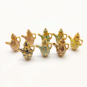Könnte beinhalten: Acht Miniatur-Teekannen aus Keramik mit goldenen Akzenten und bunten Blumenmustern. Die Teekannen sind in einem Halbkreis auf einer weißen Oberfläche angeordnet.