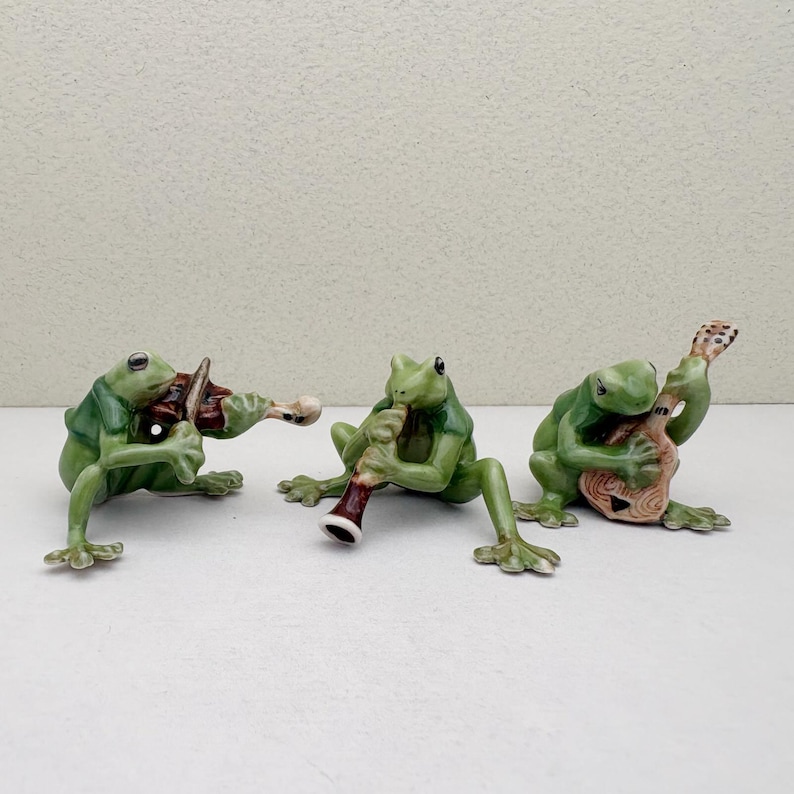 May include: Three green frog figurines playing musical instruments. One frog plays a violin, another a clarinet, and the third a guitar. The figurines are detailed with painted features and are posed as if in a band.