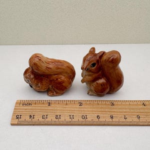 Peut inclure: Deux figurines de petits &eacute;cureuils en c&eacute;ramique marron. L'un est recroquevill&eacute;, l'autre est debout. Une r&egrave;gle en bois avec des marques en pouces est au premier plan, mesurant les figurines. Les &eacute;cureuils mesurent environ 5 cm de long.