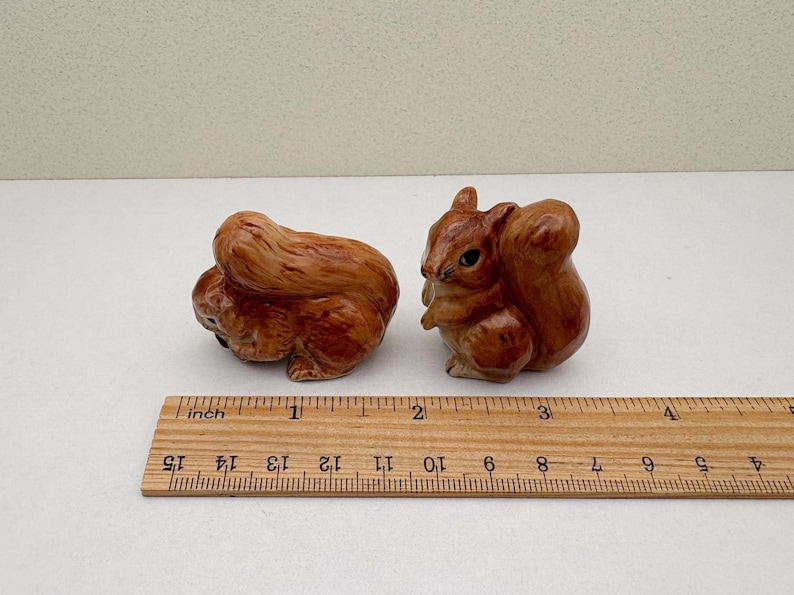 Peut inclure: Deux figurines de petits &eacute;cureuils en c&eacute;ramique marron. L'un est recroquevill&eacute;, l'autre est debout. Une r&egrave;gle en bois avec des marques en pouces est au premier plan, mesurant les figurines. Les &eacute;cureuils mesurent environ 5 cm de long.