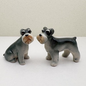 Op de afbeelding: Twee kleine keramische beeldjes van miniatuur schnauzers. Eén hond zit en de andere staat. Beide honden zijn grijs en zwart.