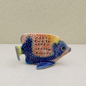 Könnte beinhalten: Eine Keramik-Fischfigur mit farbenfrohem Design. Der Fisch hat blaue, orangefarbene, gelbe und rosa Farbtöne. Die Schuppen sind detailliert, und der Fisch hat eine kleine Flosse und ein detailliertes Auge. Die Figur ist wahrscheinlich ein Dekorationsartikel.