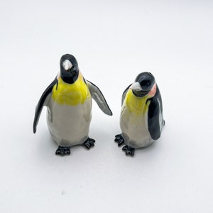 May include: Two ceramic penguin salt and pepper shakers. The penguin on the left is facing forward with its wings outstretched. The penguin on the right is facing the left with its wings tucked in. Both penguins have black and white markings and yellow beaks.