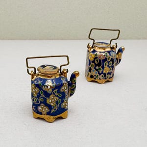 Może przedstawiać: Dwa miniaturowe czajniczki ceramiczne o głębokim niebieskim kolorze podstawy z powtarzającym się kwiatowym wzorem w kolorze złotym, białym i zielonym. Każdy czajniczek posiada uchylny uchwyt z metalu w kolorze złotym, mały dzióbek oraz pasującą pokrywkę.