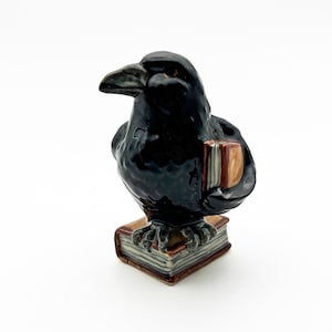 May include: A black ceramic raven figurine holding a book, perched on a stack of books. The raven has a detailed, realistic appearance.