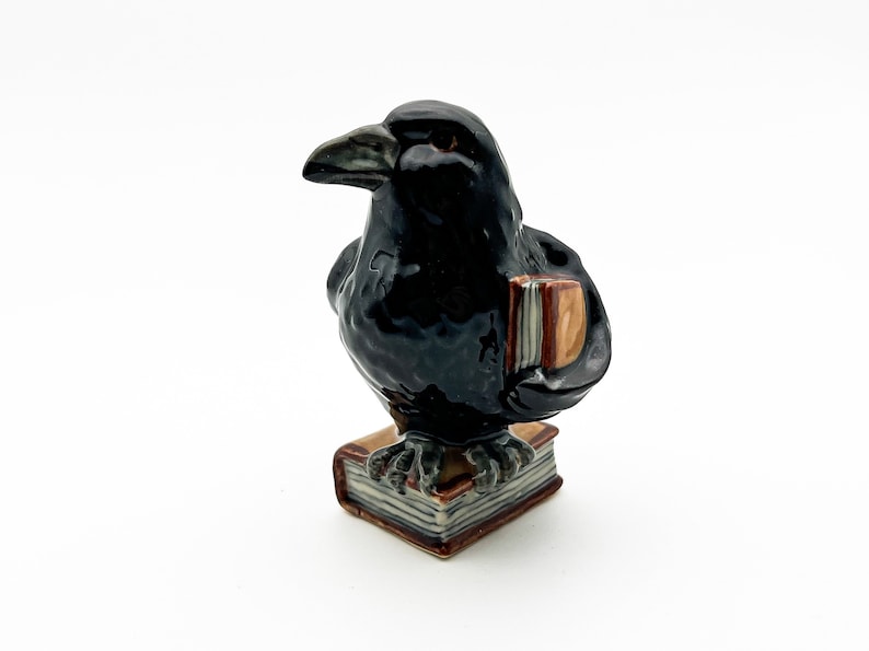 May include: A black ceramic raven figurine holding a book, perched on a stack of books. The raven has a detailed, realistic appearance.