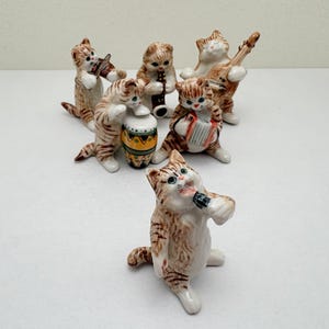 Peut inclure: Un ensemble de six figurines de chats en céramique formant un groupe de musique. Chaque chat présente des rayures tigrées brunes et joue d'un instrument différent, dont un violon, une flûte, un luth, un tambour, un accordéon et un microphone.