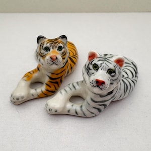 Op de afbeelding: Twee keramische tijgerbeeldjes. Eén is oranje en zwart, de andere is wit en zwart. Beide liggen in een liggende houding met hun voorpoten uitgestrekt. De tijgers hebben gedetailleerde gezichtskenmerken, waaronder groene ogen en rode neuzen.