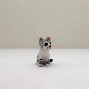 May include: A small, ceramic cat figurine. The cat is white with gray stripes and blue eyes. It is sitting upright with its tail curled. The figurine is a decorative item.