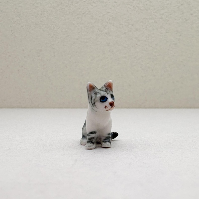 May include: A small, ceramic cat figurine. The cat is white with gray stripes and blue eyes. It is sitting upright with its tail curled. The figurine is a decorative item.