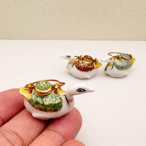 Könnte beinhalten: Drei Keramik-Entenfiguren mit dekorativen Deckeln. Die Enten sind weiß mit goldenen Akzenten und farbenfrohen Mustern auf ihren Deckeln. Eine Ente wird in einer Hand gehalten. Die Deckel weisen braune, grüne und blaue Designs auf.