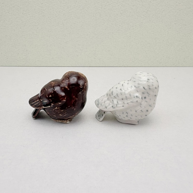 K&ouml;nnte beinhalten: Zwei Keramik-Vogelfiguren. Eine ist gl&auml;nzend dunkelbraun, die andere ist wei&szlig; mit grauen Sprenkeln. Beide befinden sich in einer Ruheposition, mit gesenktem Kopf und leicht angehobenem Schwanz. Diese dekorativen V&ouml;gel sind wahrscheinlich f&uuml;r die Inneneinrichtung gedacht.