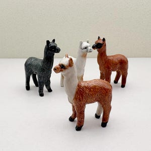 Könnte beinhalten: Vier Keramik-Alpaka-Figuren. Eine ist schwarz, eine ist weiß, eine ist braun und eine ist braun-weiß.