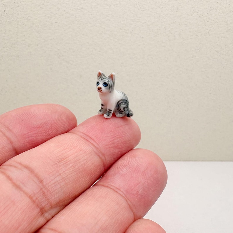 May include: A miniature ceramic kitten figurine. The small cat is white with gray stripes and blue eyes. The figurine is sitting on a finger. The cat's tail is curled. The figurine is a small decorative item.
