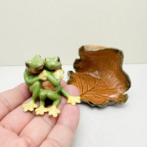Könnte beinhalten: Eine Keramikfigur von zwei grünen Fröschen, die sich umarmen, mit gelben Füßen und einem braunen Bauch. Daneben befindet sich eine braune, blattförmige Schale. Die Frösche sind der Fokus, die Schale ein ergänzendes Element.