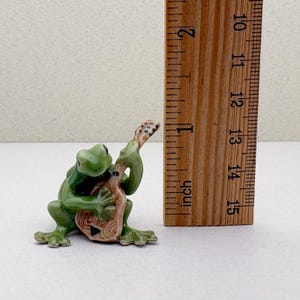 May include: A green ceramic frog figurine playing a brown guitar. The frog is approximately 1.5 inches tall. The guitar has a detailed design. The figurine is on a white surface.