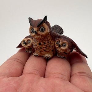 May include: A ceramic owl figurine featuring three owls. The central owl is larger, with detailed feathers and large eyes. The other two owls are smaller, with wings spread. The color palette includes brown, tan, and black.