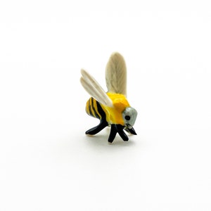 Op de afbeelding: Een klein keramiek bijenfiguurtje met zwarte en gele strepen, witte vleugels en een grijs hoofd. De bij is in een kruipende positie met zijn hoofd naar beneden.