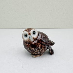 K&ouml;nnte beinhalten: Kleine Eulenfigur aus Keramik mit braun-wei&szlig; geflecktem K&ouml;rper und gro&szlig;en Augen mit schwarzen Pupillen. Die Figur steht auf einer wei&szlig;en Oberfl&auml;che, die ihre detaillierten Merkmale hervorhebt. Ein dekorativer Artikel.