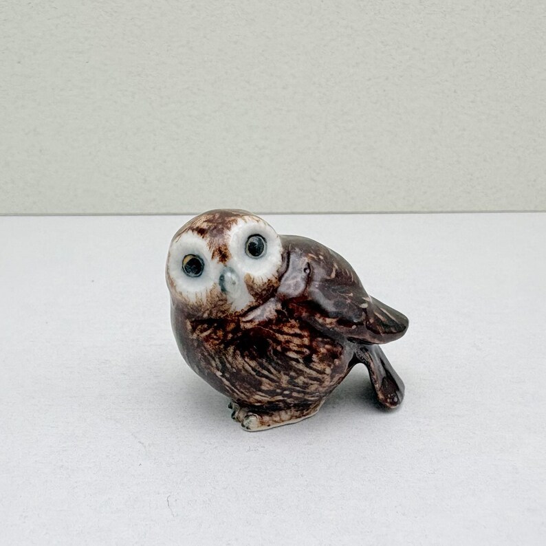 K&ouml;nnte beinhalten: Kleine Eulenfigur aus Keramik mit braun-wei&szlig; geflecktem K&ouml;rper und gro&szlig;en Augen mit schwarzen Pupillen. Die Figur steht auf einer wei&szlig;en Oberfl&auml;che, die ihre detaillierten Merkmale hervorhebt. Ein dekorativer Artikel.