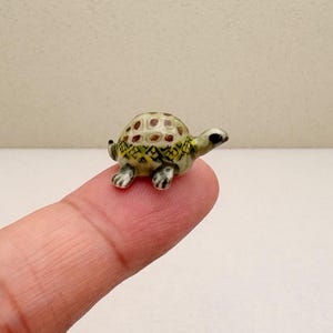 Może przedstawiać: Mała, ceramiczna figurka żółwia z zielono-brązowym pancerzem. Żółw idzie po palcu.
