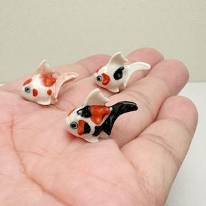 May include: Three miniature koi fish figurines. The ceramic fish have white bodies with orange, black, and red markings. The fish are displayed on a light-colored surface.
