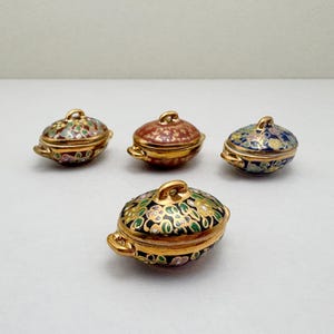 May include: Four miniature oval-shaped lidded tureens with gold-tone handles and rims. Each piece features intricate floral enamel patterns in distinct color combinations including black, blue, red, and multi-colored designs with gold accents.