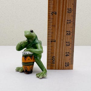 May include: A green ceramic frog figurine playing a small drum. The drum is decorated with orange and black patterns. The figurine is approximately 2 inches tall.