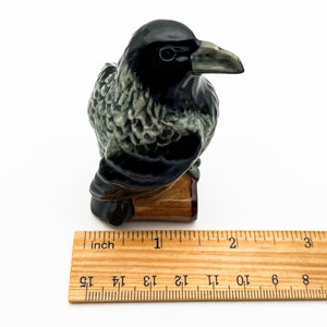 May include: A black and green ceramic crow figurine perched on a wooden base. The crow is about 2 inches tall.